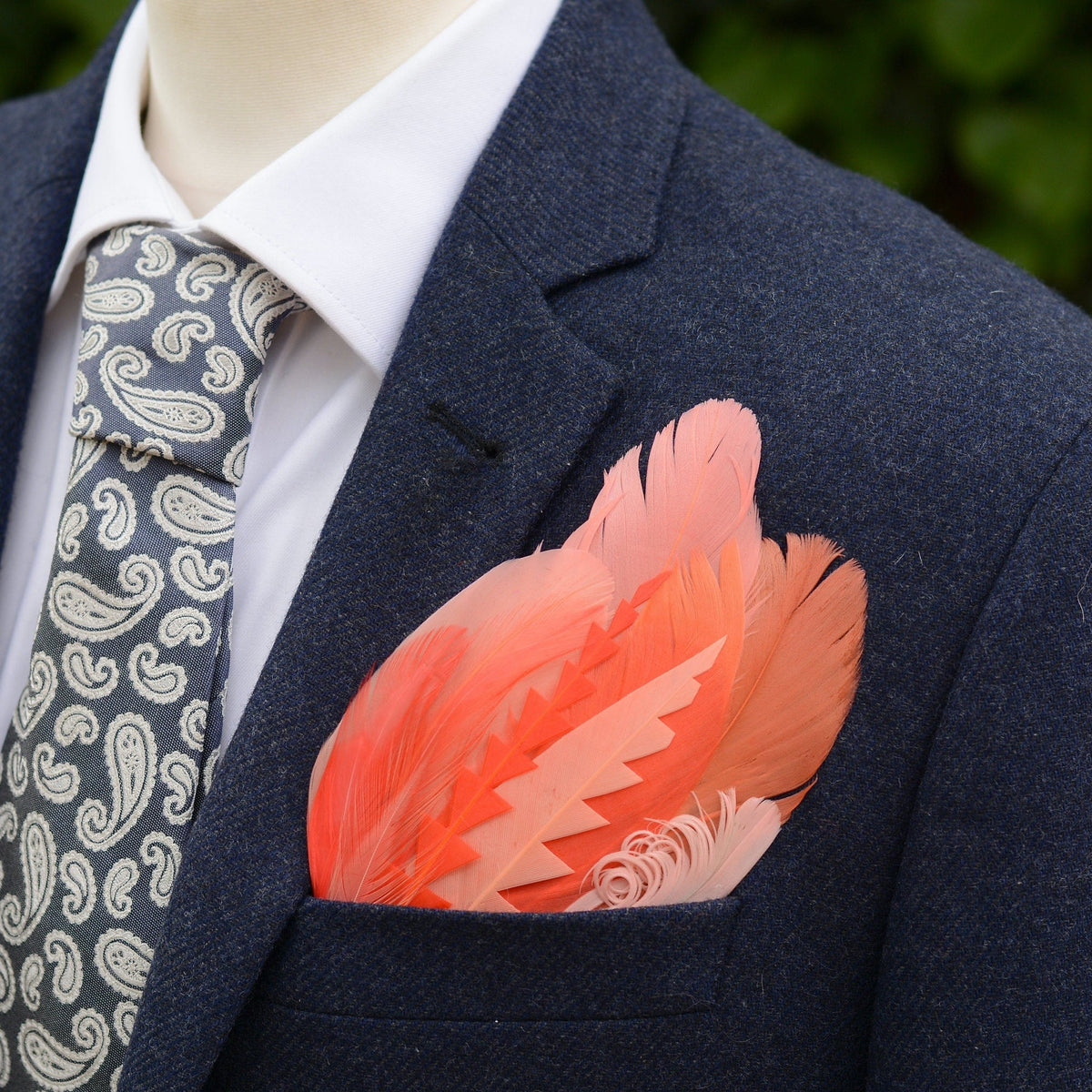 Blue Tweed suit jacket with a large tropical-style feather pocket square in shades of coral and pink laid on a grey stone.