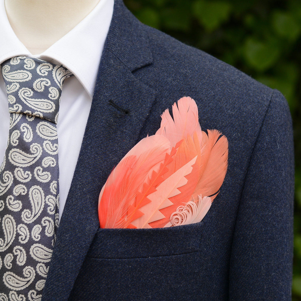 Blue Tweed suit jacket with a large tropical-style feather pocket square in shades of coral and pink laid on a grey stone.