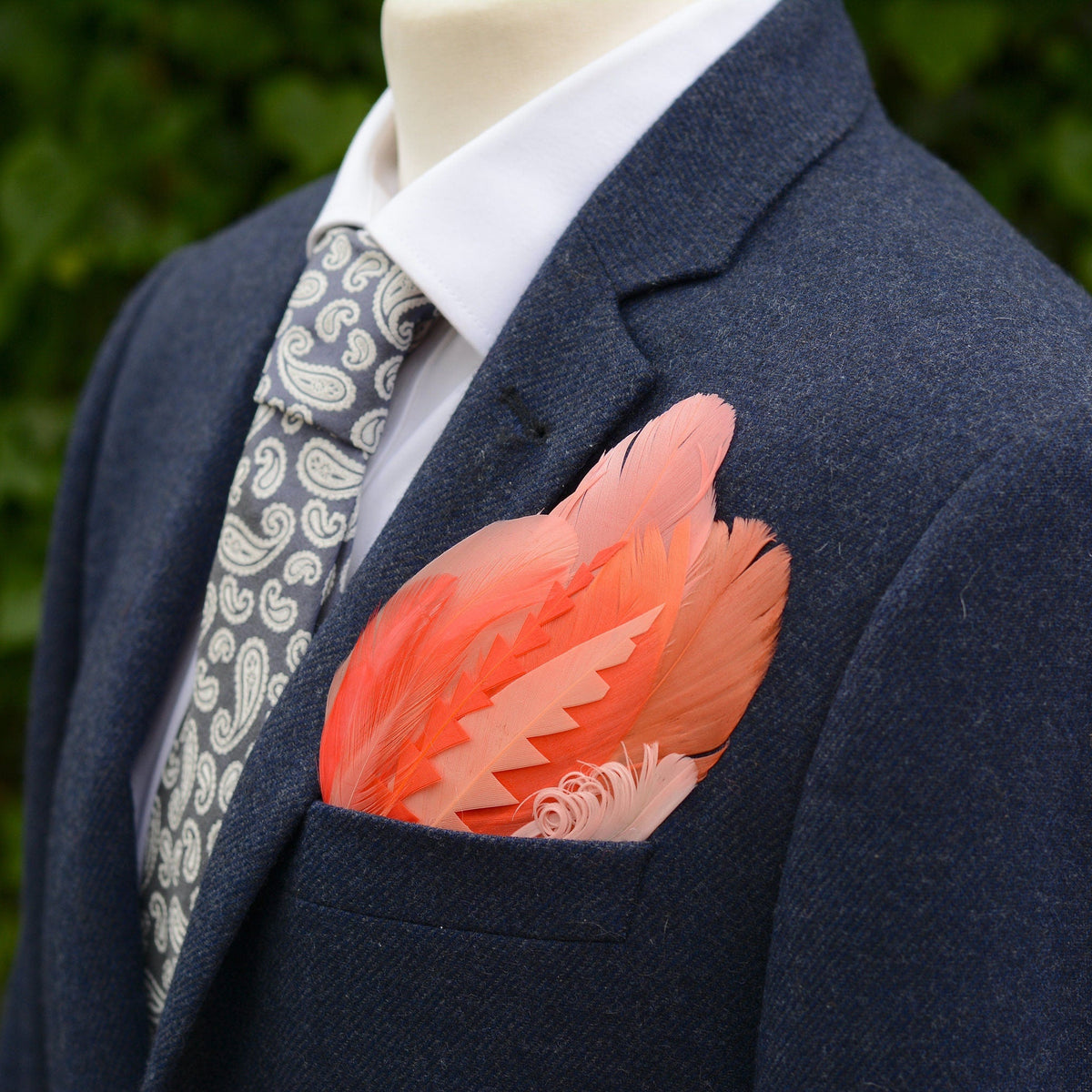 Blue Tweed suit jacket with a large tropical-style feather pocket square in shades of coral and pink laid on a grey stone.