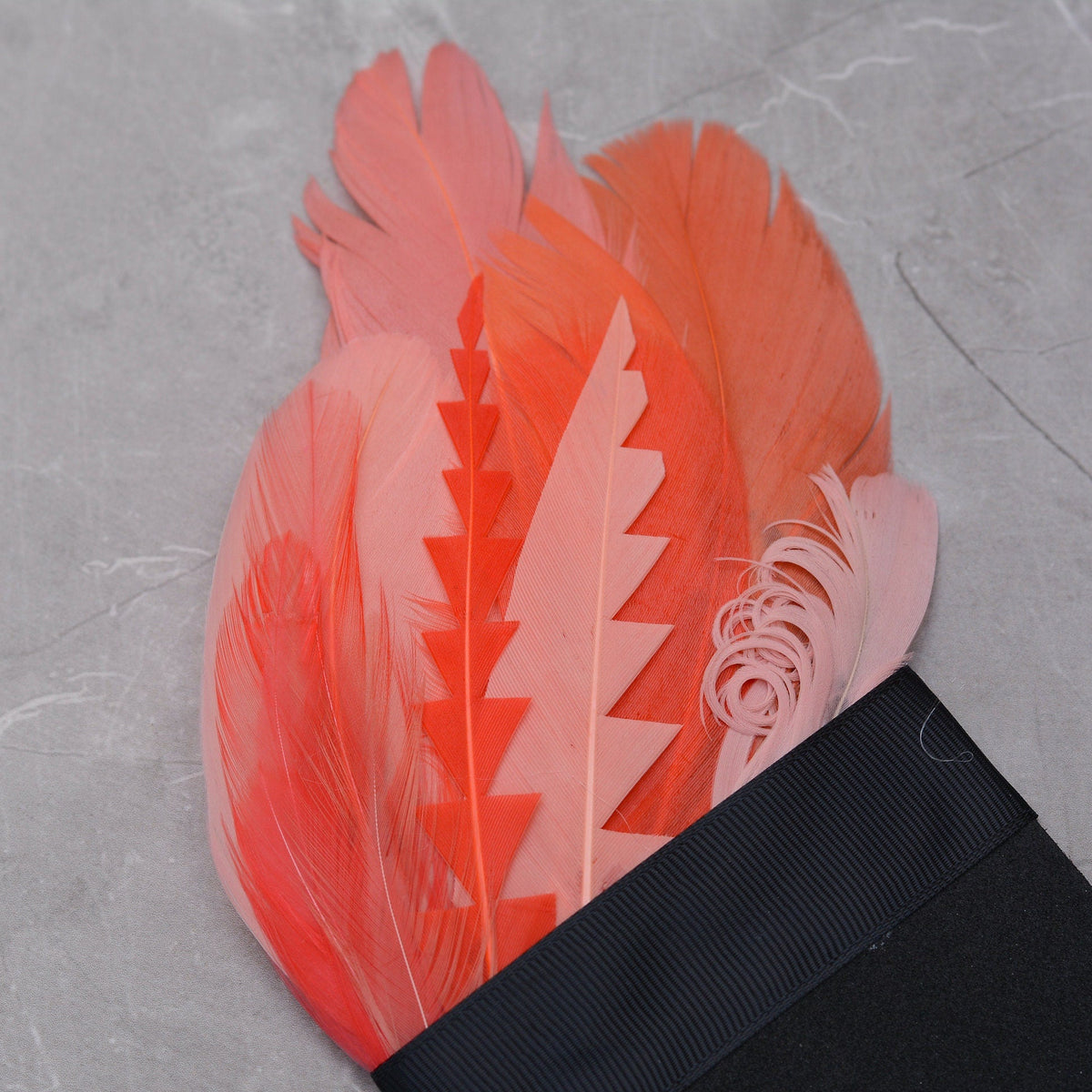Close up of a tropical-style feather pocket square in shades of coral and pink laid on a grey stone.