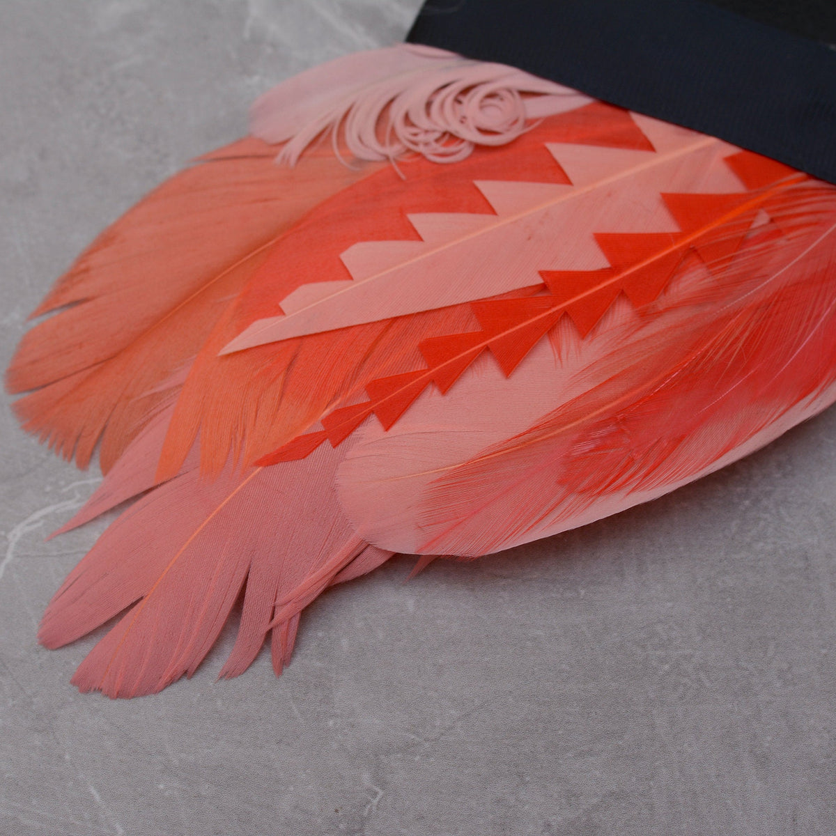 Close up of a tropical-style feather pocket square in shades of coral and pink laid on a grey stone.