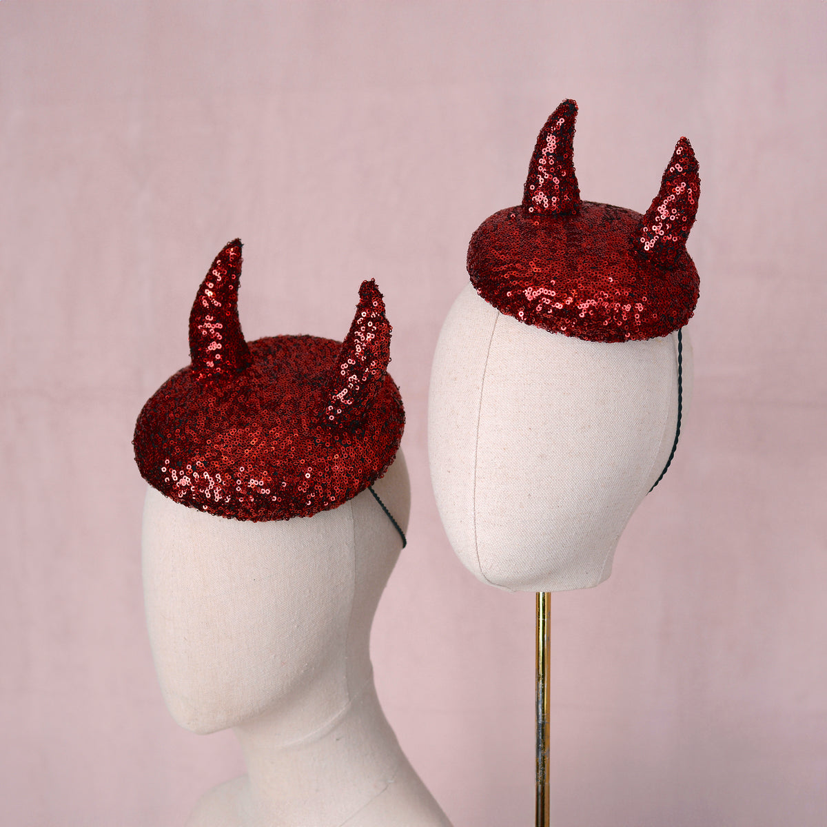 Two red sequin devil horn hats displayed on white mannequin heads against a soft pink background.