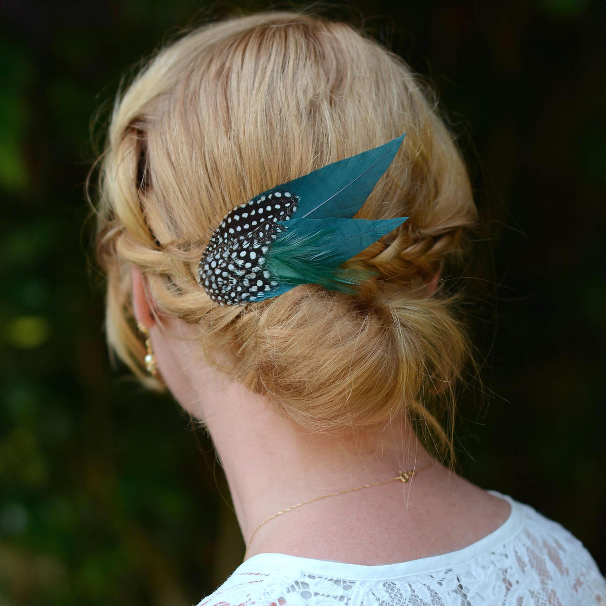 A blonde woman shown from the back wearing a dark teal green and black polka dot feather hair accessory in her braided hair, with a white lace top and a blurred dark green background.