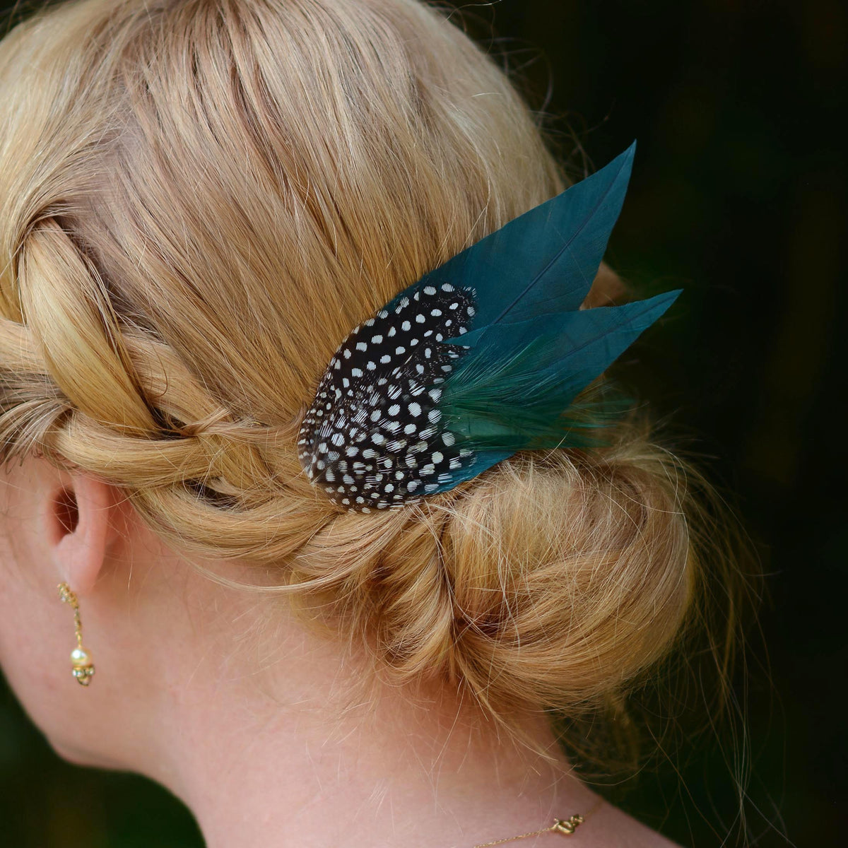 A close-up side view of a blonde woman's hair styled in a low braided bun, featuring a decorative hair accessory with deep teal green feathers and a black feathers  covered in white spots. An ideal hair clip for formal events and weddings.