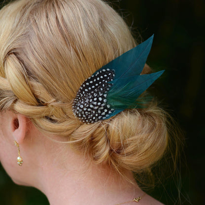 A close-up side view of a blonde woman's hair styled in a low braided bun, featuring a decorative hair accessory with deep teal green feathers and a black feathers  covered in white spots. An ideal hair clip for formal events and weddings.