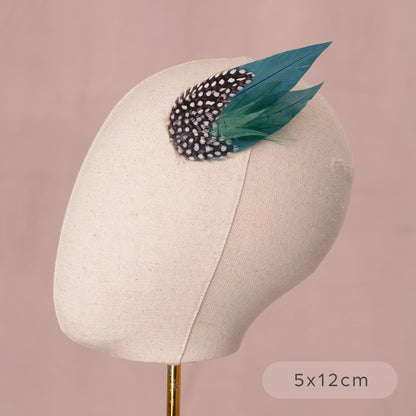 A feather hair clip fascinator in deep teal green and polka-dot spotted feathers. Displayed on a cream canvas mannequin against a soft pink background.