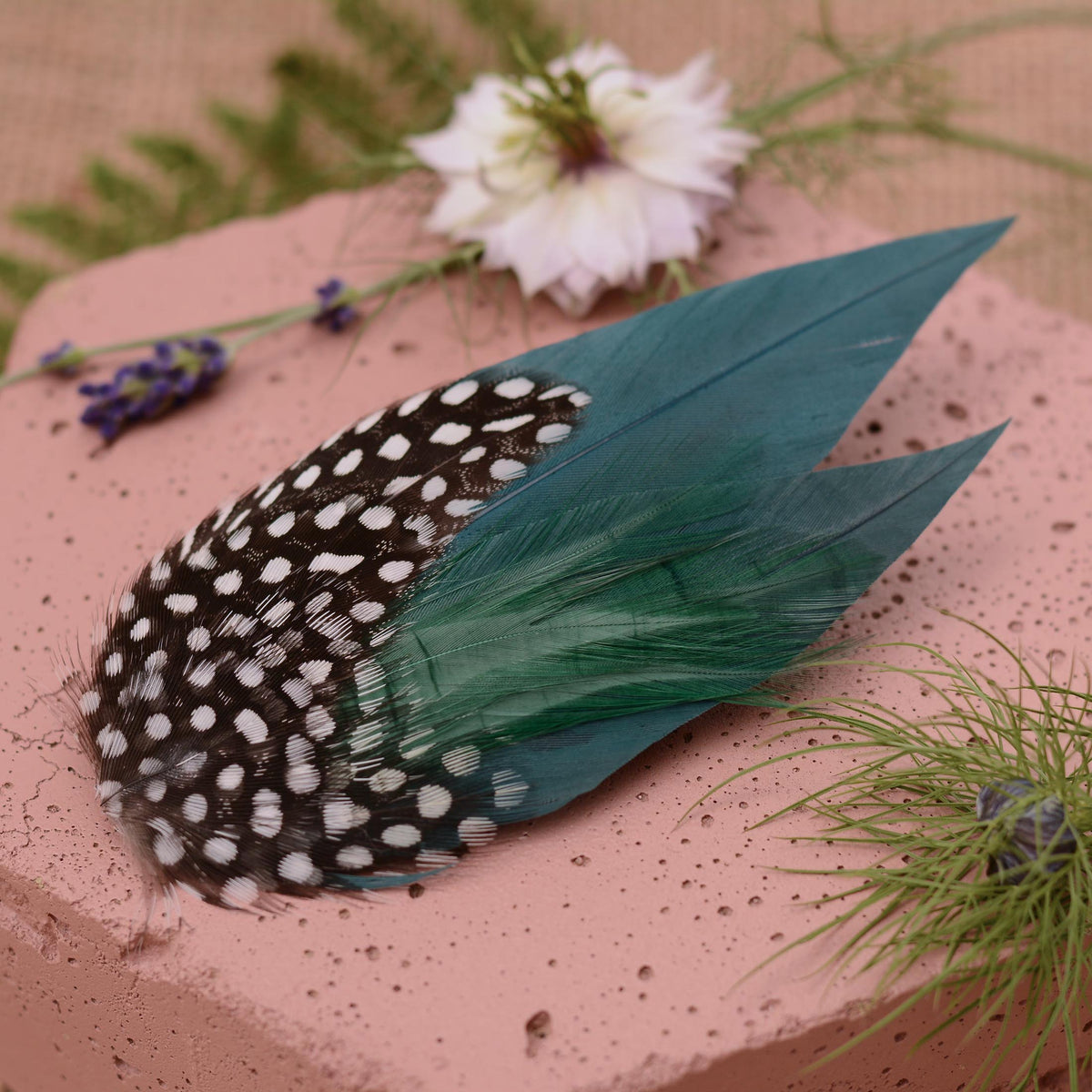 A decorative feather hair accessory with black and white spotted feathers on  deep teal green feathers, styled on a pink textured surface with dried flowers and greenery.