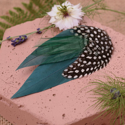 A decorative feather hair clip in deep teal green and black and white polka dot feathers. On a pink stone with flowers and greenery around it.