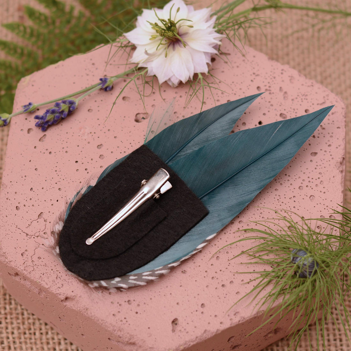 A decorative hair  clip featuring a black felt back and silver tone alligator clip cushioned with felt, displayed on a pink textured stone with floral props.