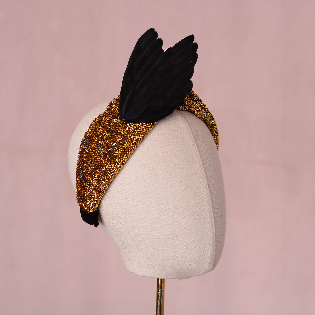 A black bird wing feather fascinator headpiece with a gold glitter fabric headband base, displayed on a cream fabric mannequin head against a soft pink background.