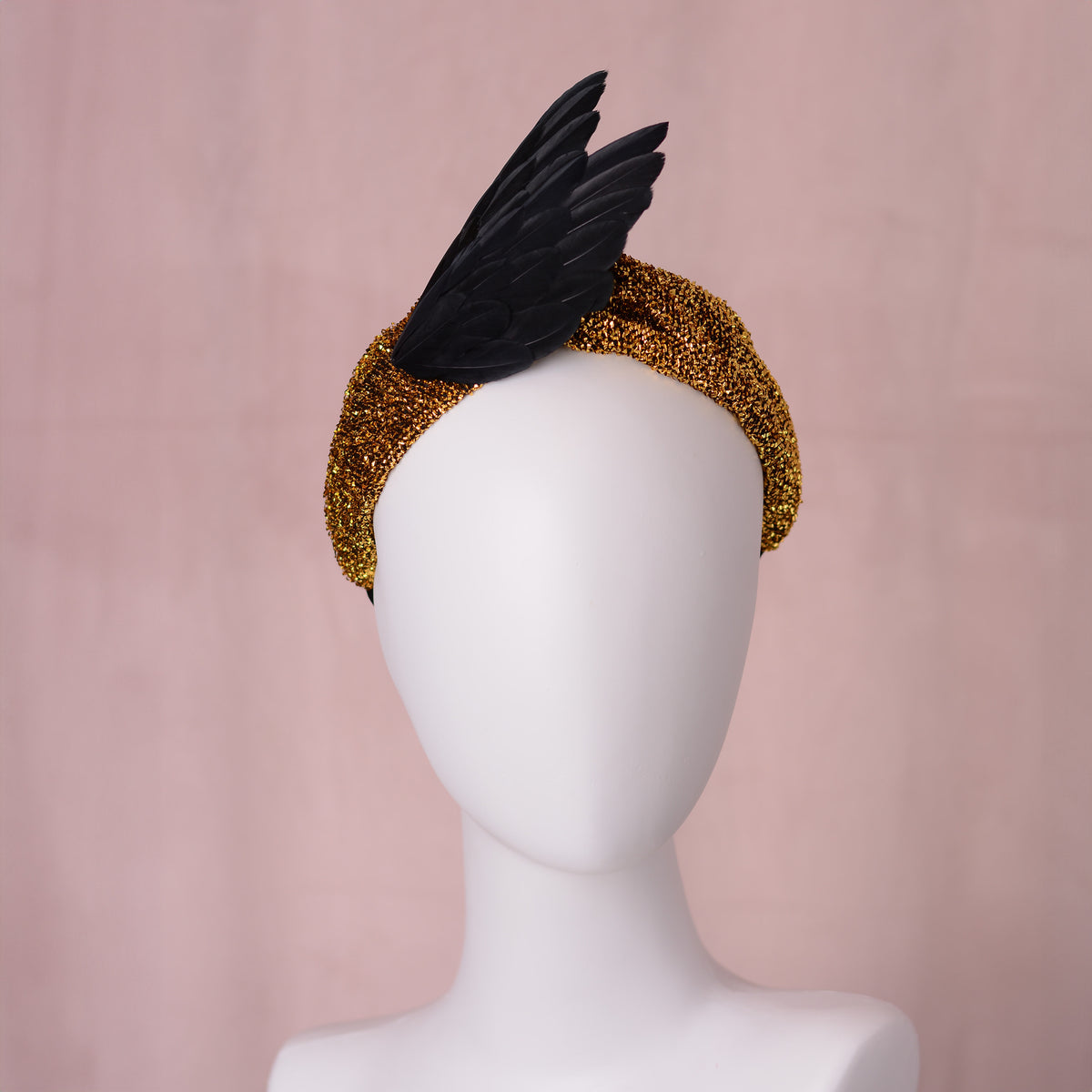 A black feather fascinator on a sparkling gold glitter turban headband, displayed on a white mannequin head against a soft pink backdrop. It is giving witchy fortune teller vibes