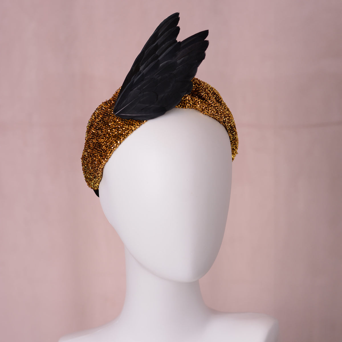 A black bird wing feather fascinator headpiece with a textured gold glitter head band, displayed on a white mannequin head against a soft pink backdrop.