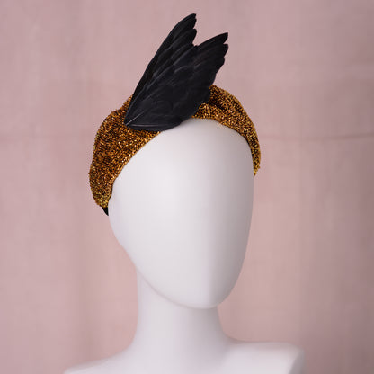 A black bird wing feather fascinator headpiece with a textured gold glitter head band, displayed on a white mannequin head against a soft pink backdrop.