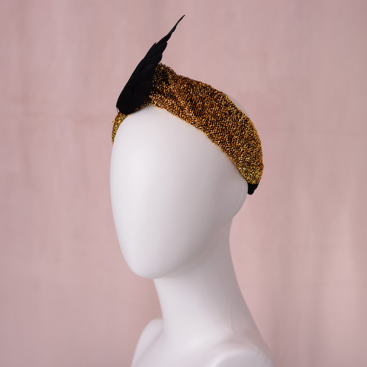 A black winged feather fascinator with a textured gold glitter band, displayed on a white mannequin head against a soft pink background.