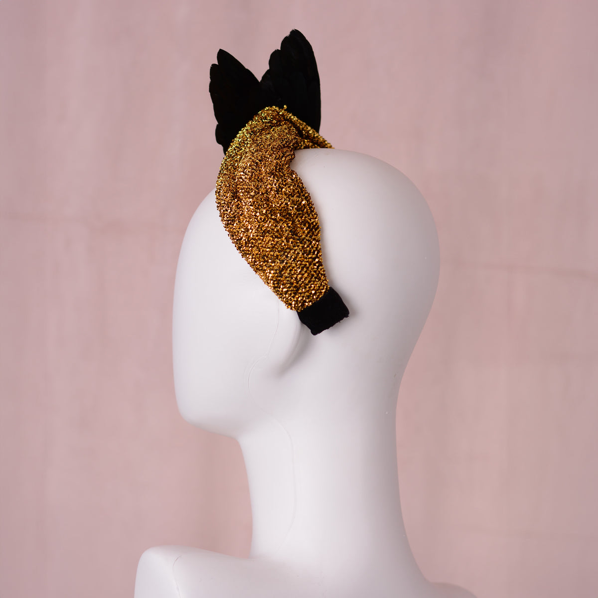 A gold glitter headband with black feather wings, displayed on a white mannequin head against a soft pink background.