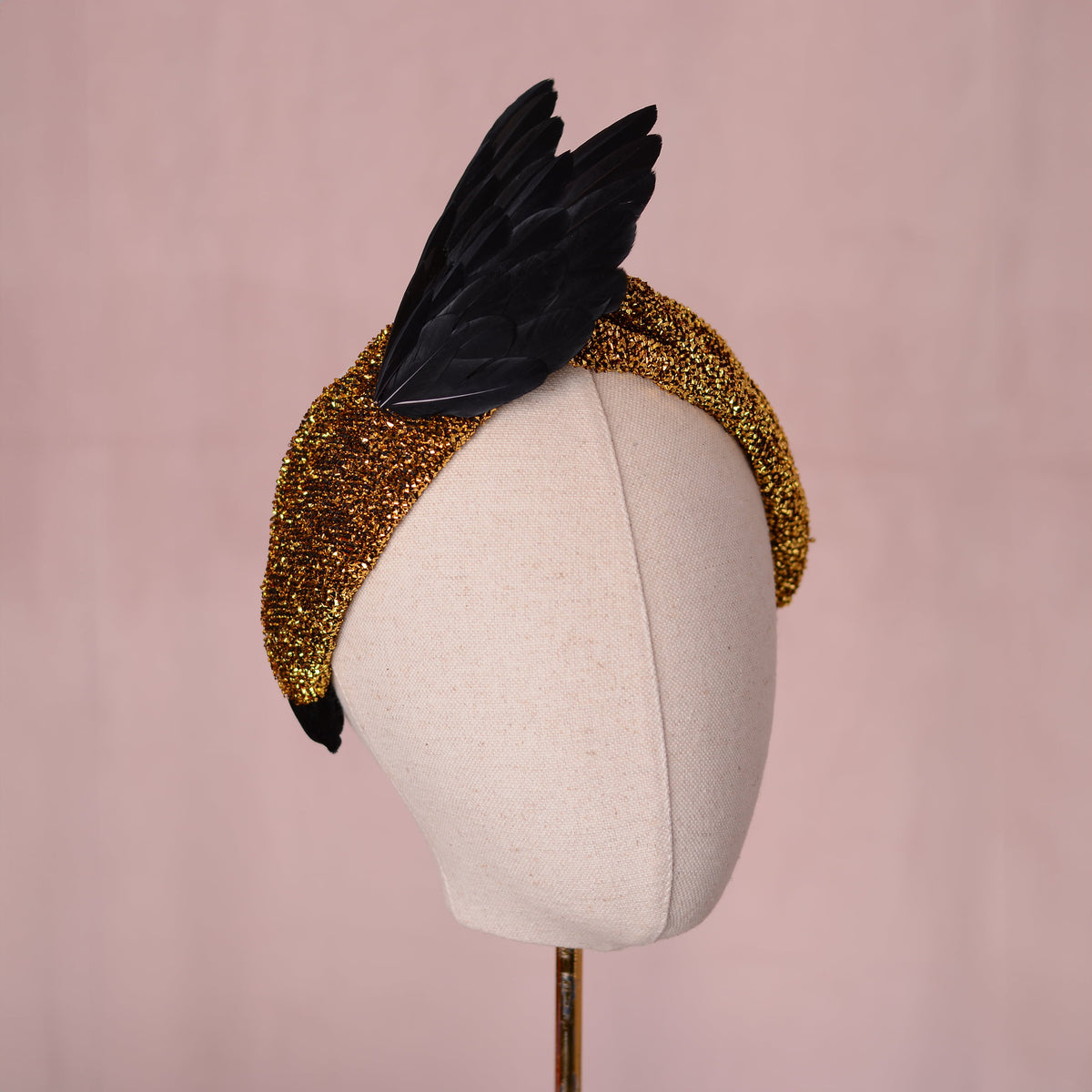 A gold fascinator headpiece with black feathers in a wing design, displayed on a cream fabric mannequin head against a soft pink background.