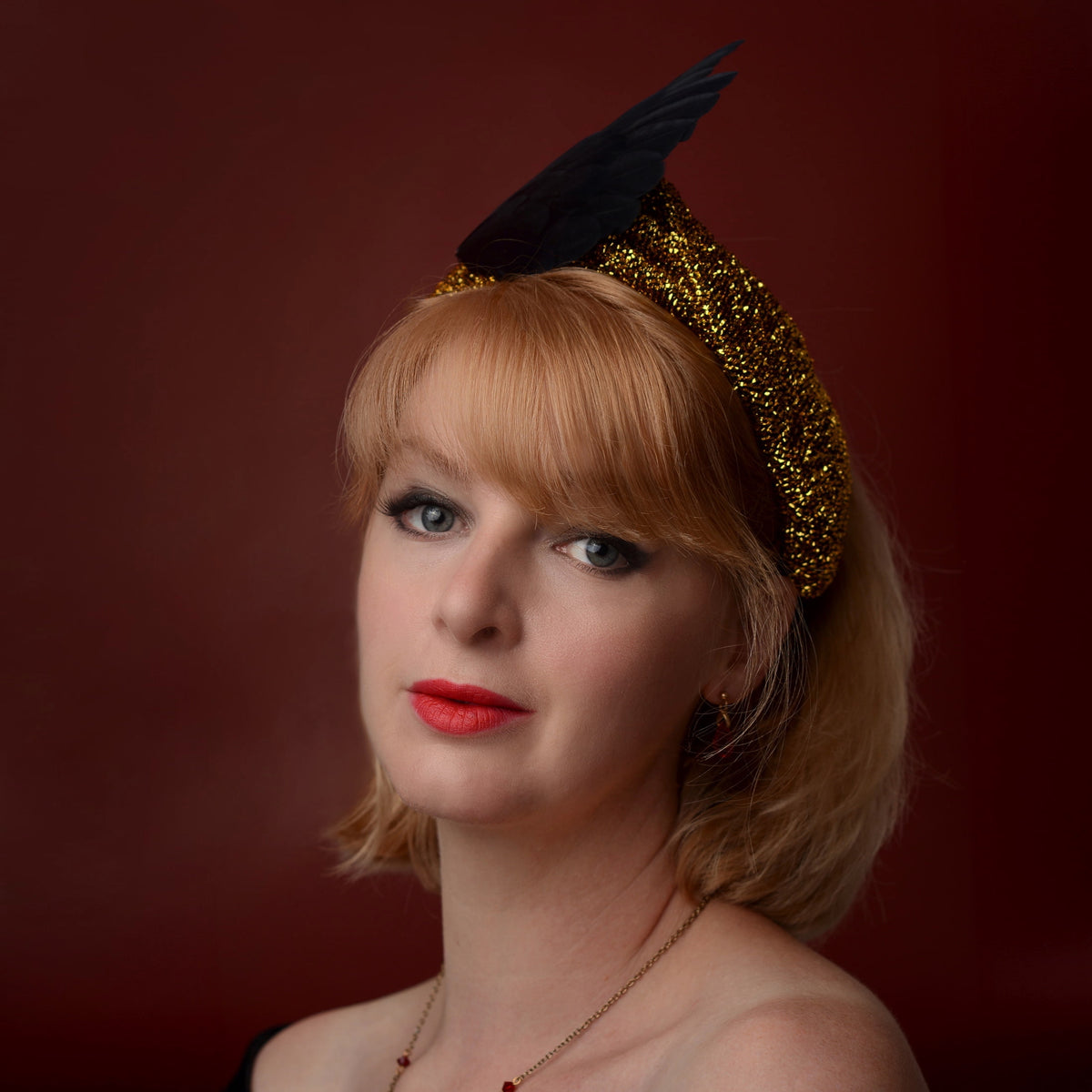 A woman with short blonde hair and red lipstick wears a black winged feather fascinator headband with a gold glitter band, posed against a dark red studio background.