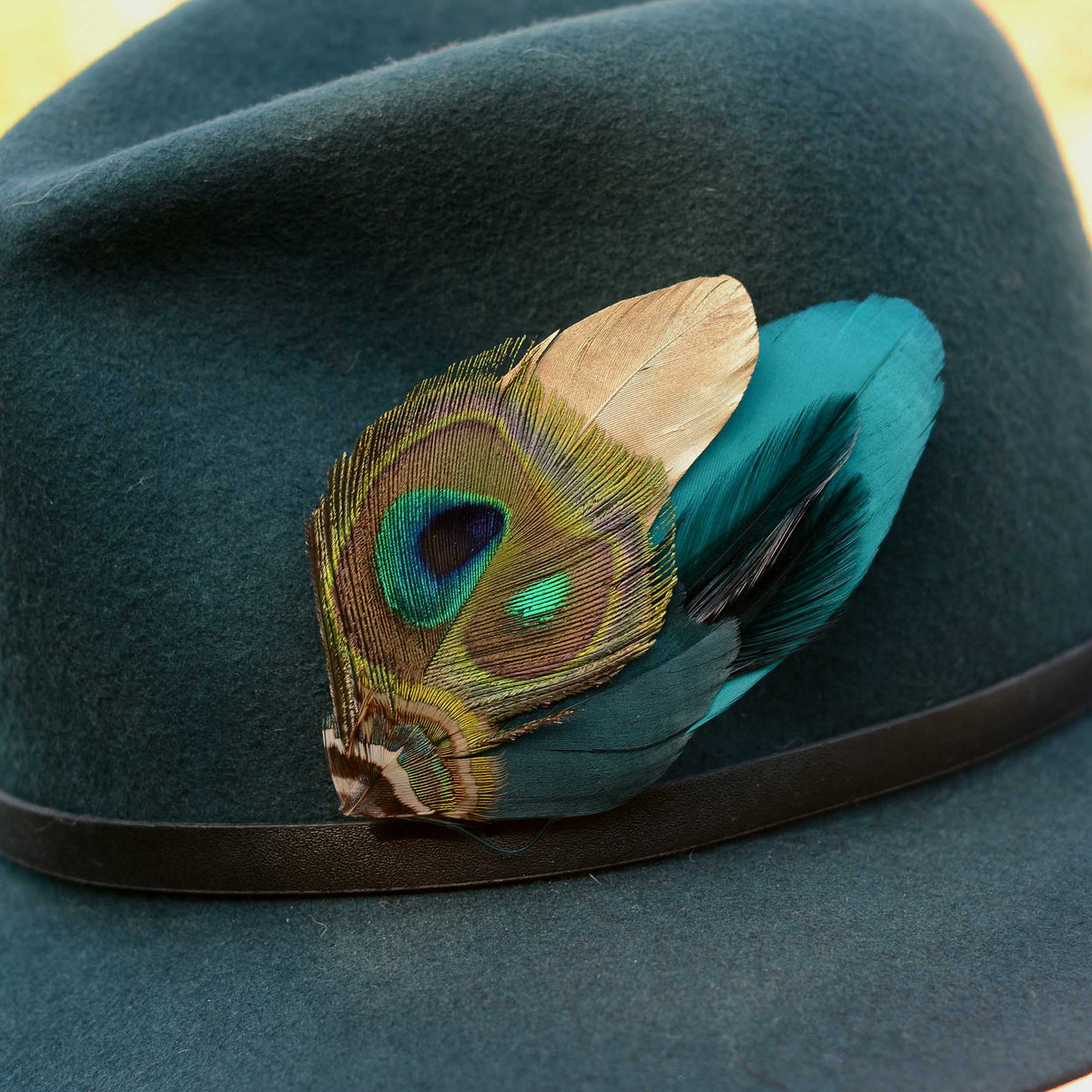 Close up of a teal fedora hat with a gold and teal peacock feather hat pin, placed on a metal bird stand against an outdoor blurred background
