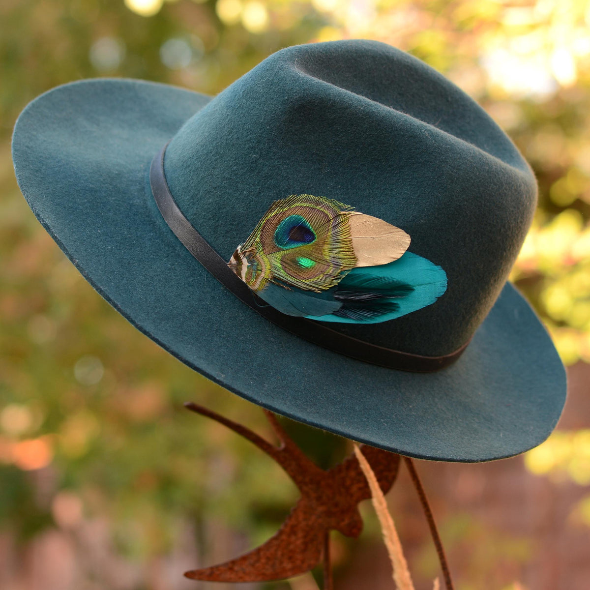 Teal fedora hat with a gold and teal peacock feather hat pin, placed on a metal bird stand against an outdoor blurred background