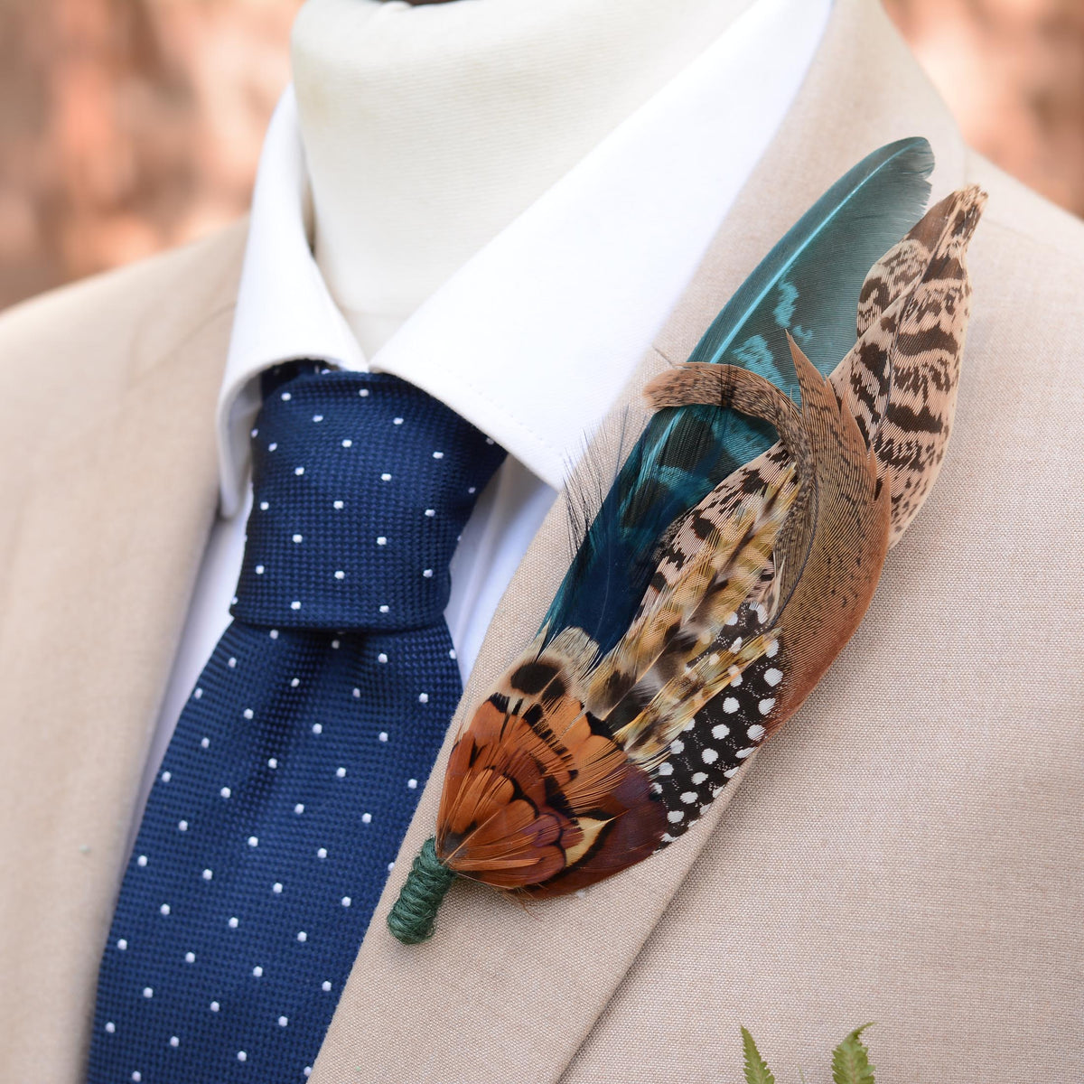 A close-up of a formal outfit featuring a beige suit jacket, white dress shirt, and a navy blue polka-dot tie, accessorised with a large, decorative feathered boutonniere on the lapel in natural pheasant feathers paired with a green dyed wing feather.