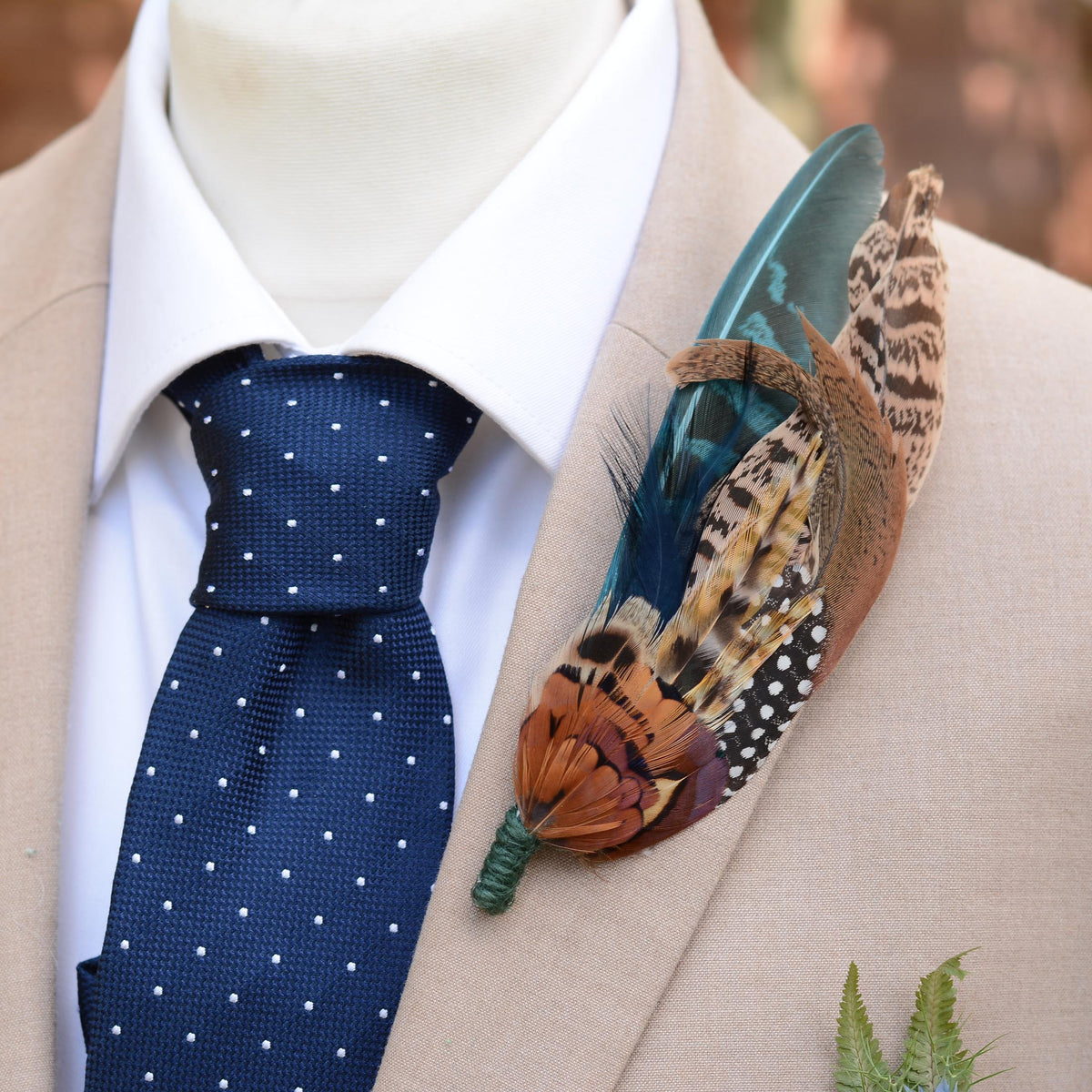 A decorative feathered wedding boutonniere pin displayed on a beige suit jacket and white dress shirt with a navy polka dot tie. The pin features a teal blue tail feather, a brown and black spotted patterned feather, and warm orange-brown feathers with a green wrapped stem.