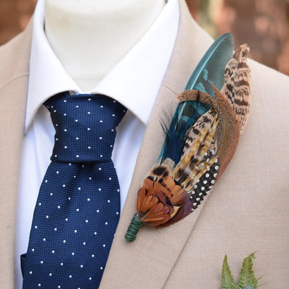 A decorative feathered wedding boutonniere pin displayed on a beige suit jacket and white dress shirt with a navy polka dot tie. The pin features a teal blue tail feather, a brown and black spotted patterned feather, and warm orange-brown feathers with a green wrapped stem.