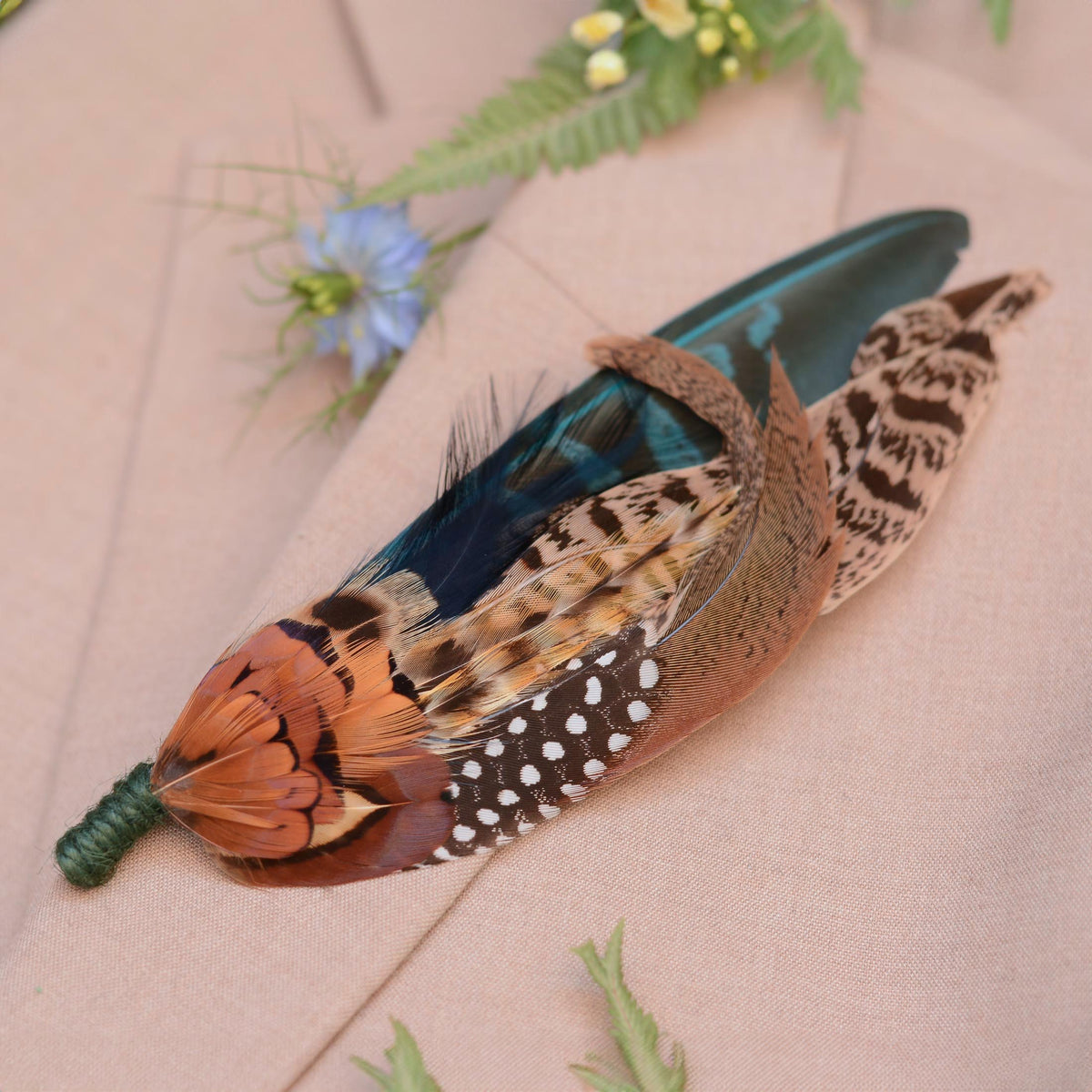 A decorative feather brooch pin featuring a vibrant orange-brown patterned pheasant feathers, an teal feather, and a brown-and-white spotted feather, displayed on a beige fabric background with soft green and blue floral accents.