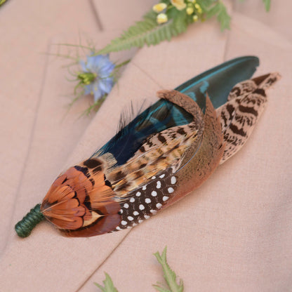 A decorative feather brooch pin featuring a vibrant orange-brown patterned pheasant feathers, an teal feather, and a brown-and-white spotted feather, displayed on a beige fabric background with soft green and blue floral accents.
