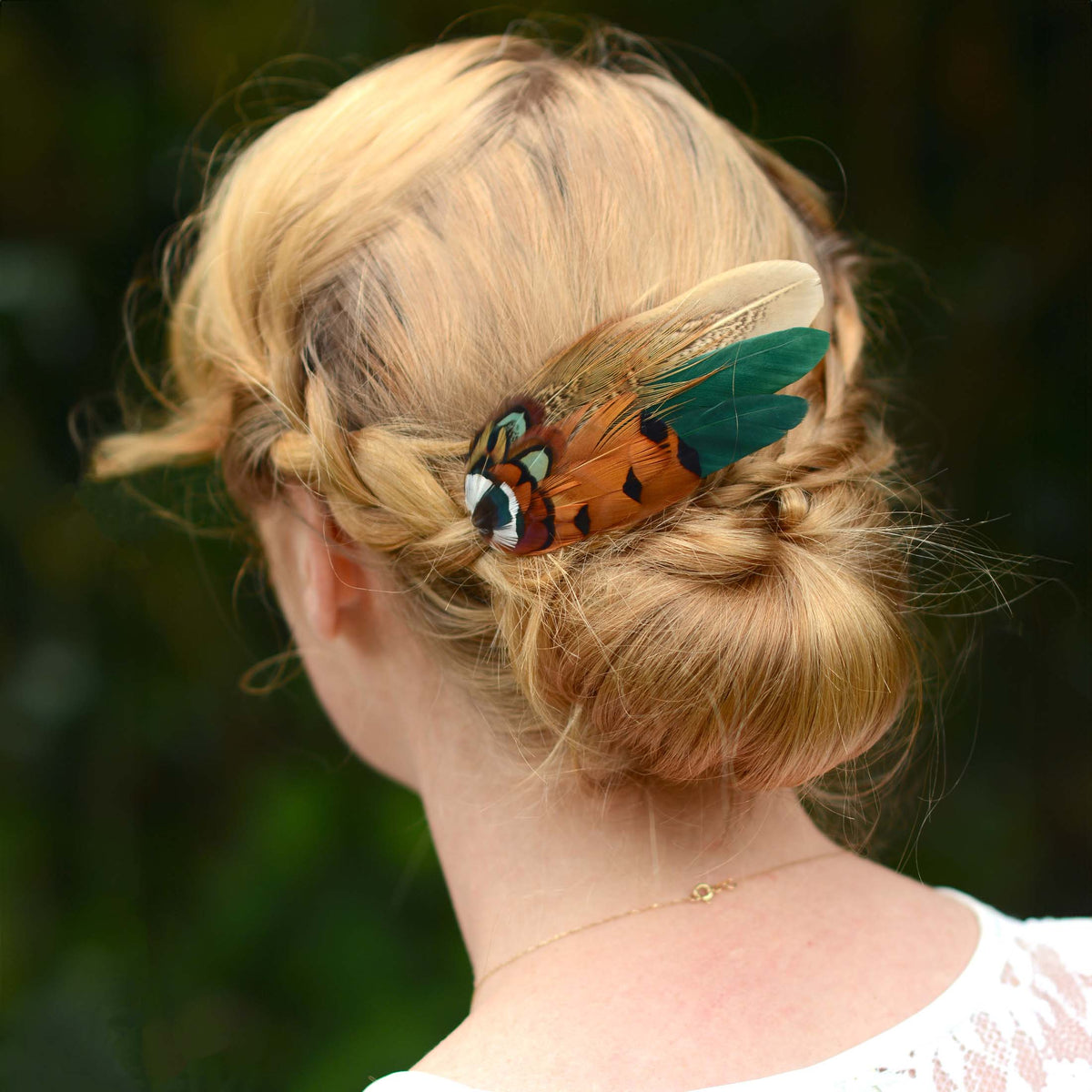 A blonde woman shown from the back wearing a decorative pheasant feather hair clip fascinator in her low bun, with a blurred green outdoor background.