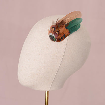 A decorative pheasant feather hair accessory with copper, brown and green feathers arranged in a wing-like design displayed on a cream fabric mannequin head against a soft pink background.