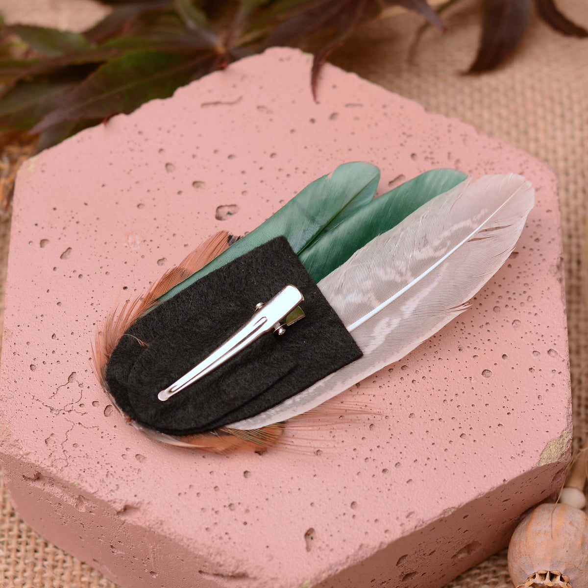 A feather hair clip with a black felt base, and silver alligator-style hair clip resting on a pink textured stone block.