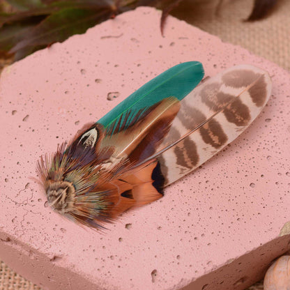 A decorative feather hair fascinator with a green feather, and natural pheasant feathers in copper, brown and green, displayed on a pink stone slab.