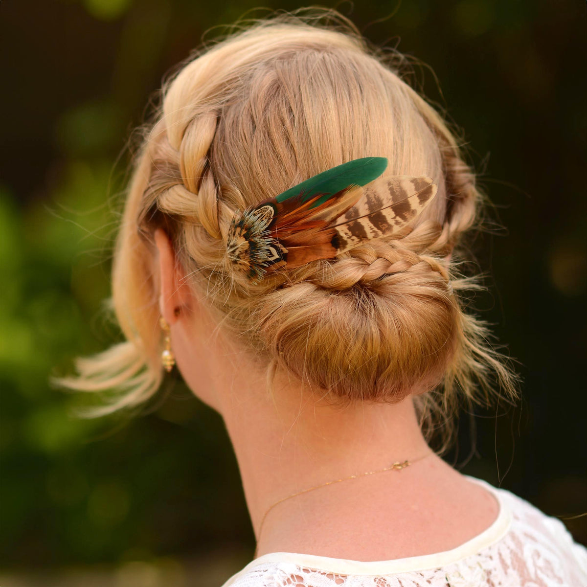 A blonde woman shown from the back wearing a braided low bun with a decorative feather hair accessory in green, brown, and black.