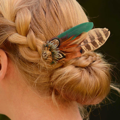 A close-up of a blonde woman's braided hair bun decorated with a feather hair accessory featuring a green feather, and patterned pheasant feathers.