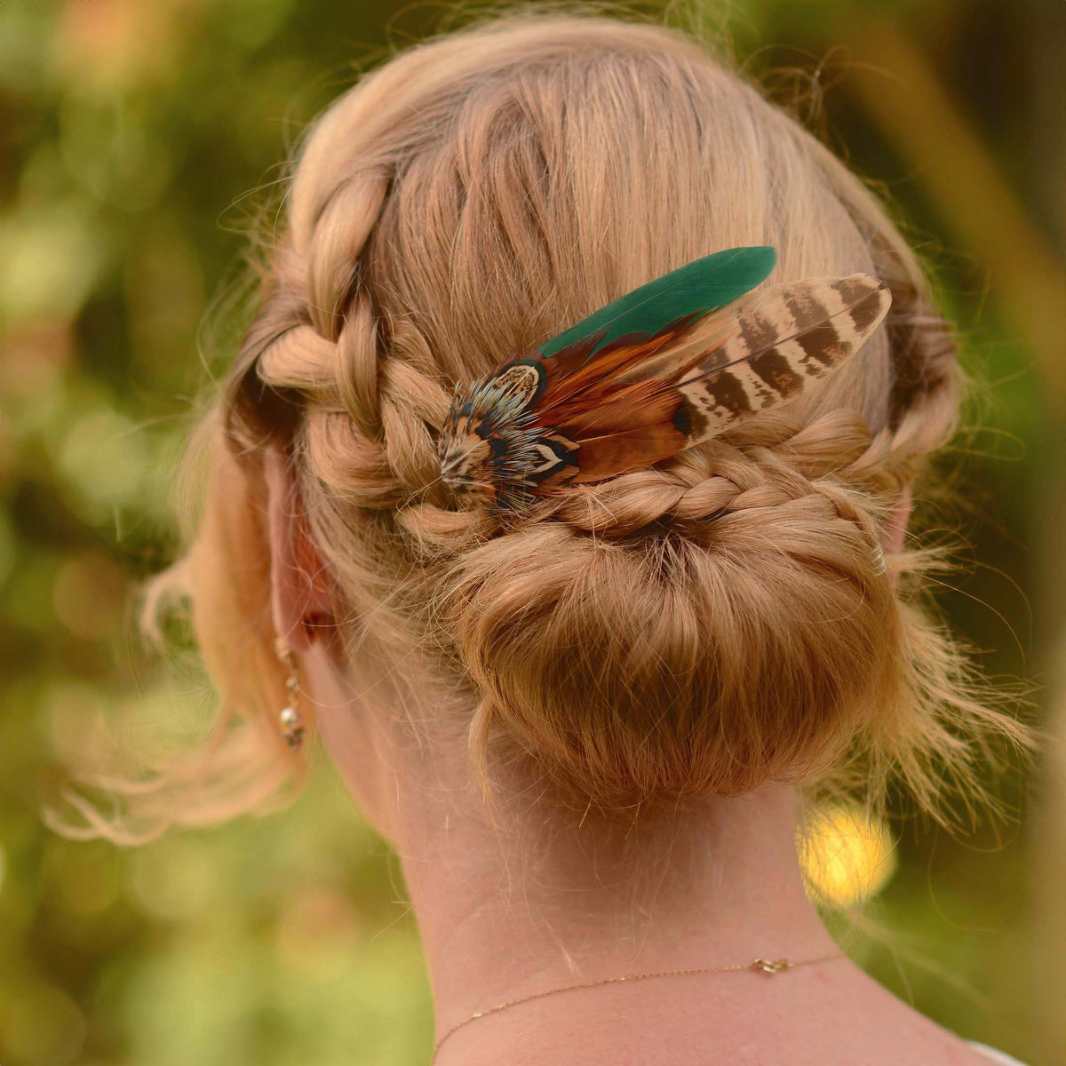 A blonde woman shown from the back wearing a braided updo hairstyle with a decorative pheasant feather hair accessory in green, brown, and black.
