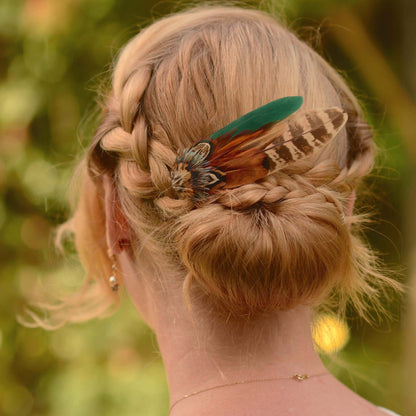 A blonde woman shown from the back wearing a braided updo hairstyle with a decorative pheasant feather hair accessory in green, brown, and black.