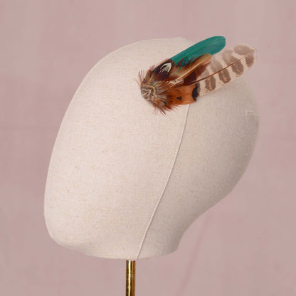 A decorative feather accessory with brown patterned feathers and a green accent feather, displayed on a cream fabric mannequin head against a soft pink background.