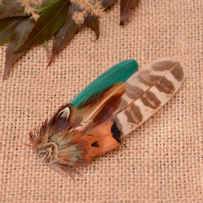 A decorative feather fascinator in natural pheasant feathers in copper, black, brown and cream and a bottle green duck feather in a wing shape, displayed on a beige burlap surface with dried leaves in the corner. 