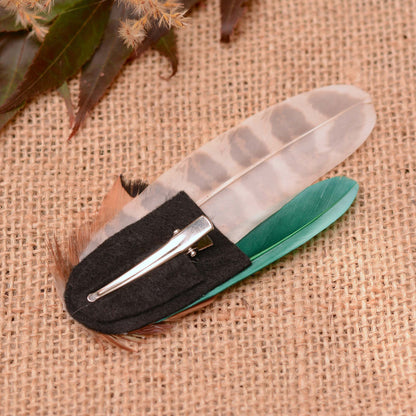 A pheasant feather hair clip placed upside down to show the black felt base and silver alligator clip, displayed on a burlap background.