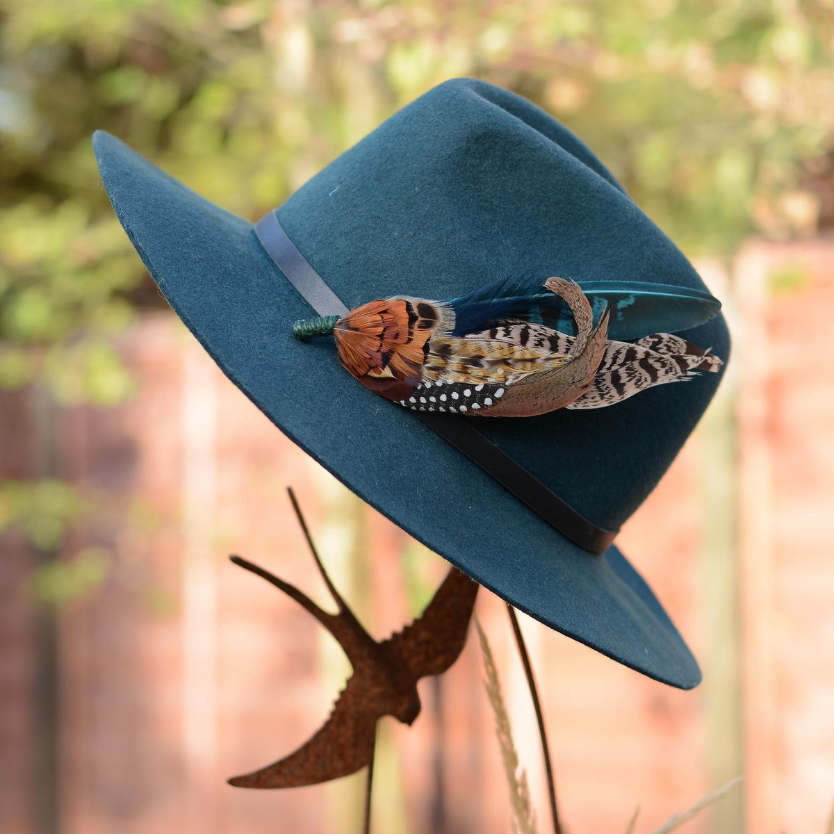 A teal blue wide-brim fedora hat with a black band, decorated with a pheasant feather  hat pin, displayed outdoors against a blurred fence and foliage background.