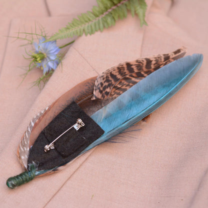 A decorative feather pin with a turquoise blue feather, brown and black patterned feather, and a black felt backing with a silver safety pin, displayed on a beige fabric background with a small blue flower and green fern.