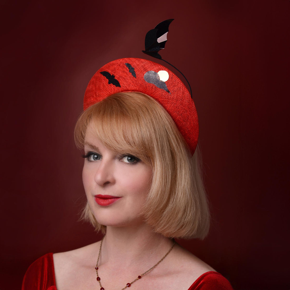 A woman with a short blonde bob wears a bright red fascinator hat decorated with black bat silhouettes, a cloud, and a metallic pink blood moon with a floating bat, posed against a dark red background.
a headpiece for Halloween-themed weddings, events and parties