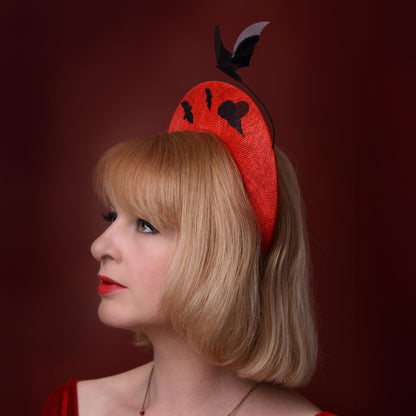 A woman in profile wearing a bright red fascinator hat with black sequin bat and cloud motifs and a floating bat accent, set against a dark red studio background. A headpiece for special Halloween-themed events and parties.