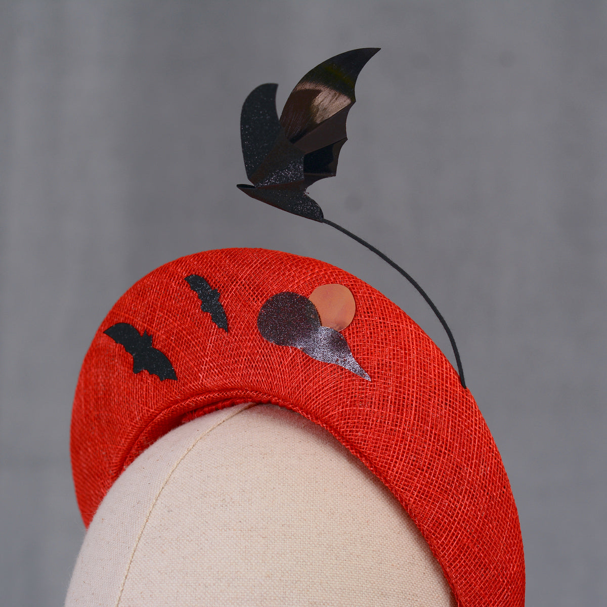 A red Halloween headband featuring black bat and moon decorations. The headband has a textured finish and is decorated with two bat silhouettes and a moon design and a larger  floating bat.