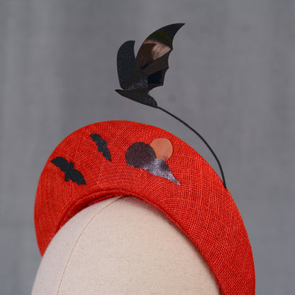 A red Halloween headband featuring black bat and moon decorations. The headband has a textured finish and is decorated with two bat silhouettes and a moon design and a larger  floating bat.