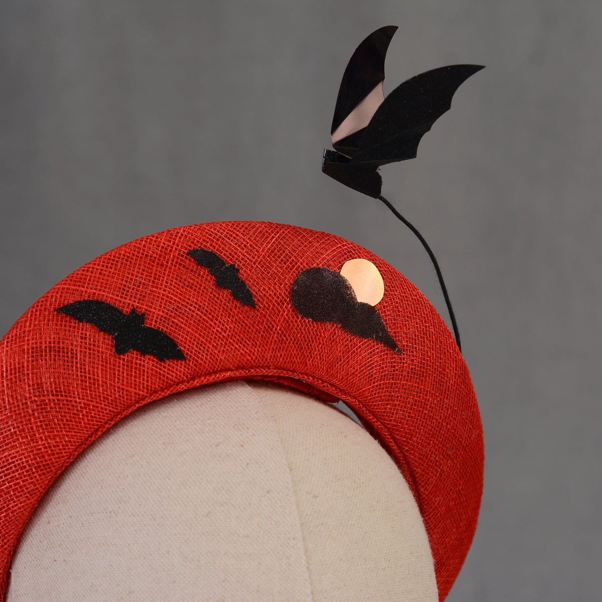 A red Halloween headband featuring black bat and moon decorations. The headband has a textured finish and is decorated with two bat silhouettes and a moon design and a larger  floating bat.