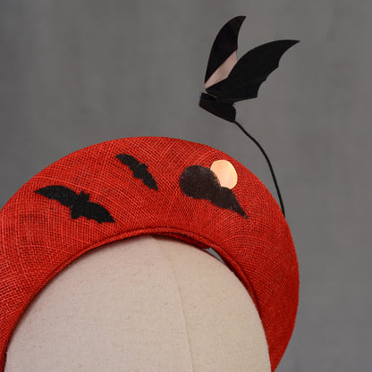 A red Halloween headband featuring black bat and moon decorations. The headband has a textured finish and is decorated with two bat silhouettes and a moon design and a larger  floating bat.
