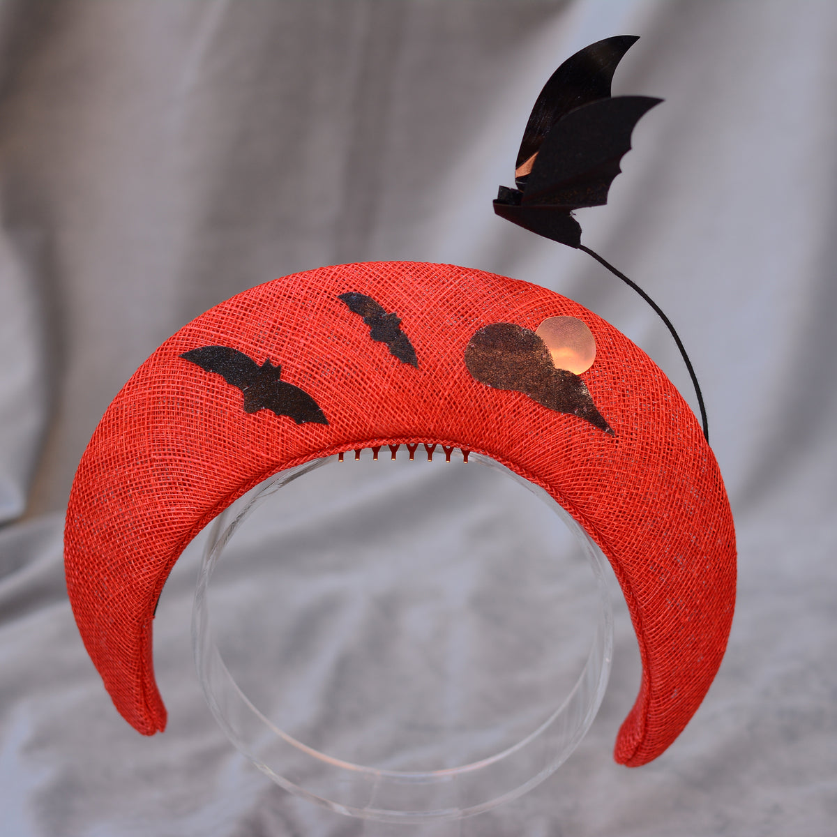 A vibrant red headband with a Halloween design. It features black bat and cloud motifs, with a black bat accent extending upwards. The headband is made of a textured straw and is mounted on a clear stand.
