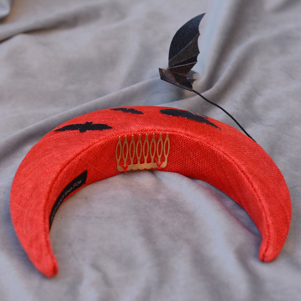 A red crescent-shaped headpiece with black bat silhouettes and a small, floating bat. The headpiece has a gold comb for securing it in the hair. The fabric is a textured straw.
