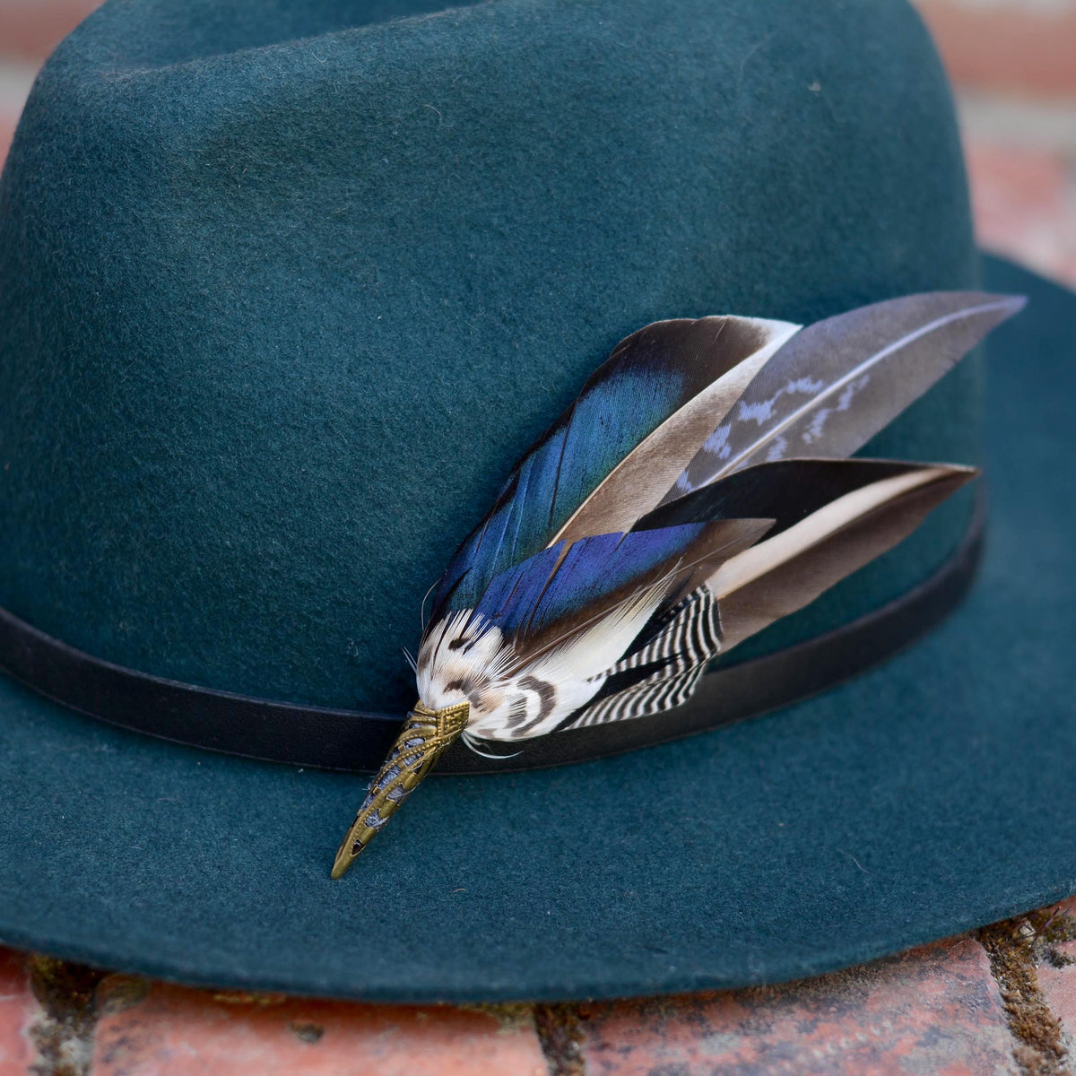 A  close up of a iridescent blue mallard duck wing feather hat pin  on a teal fedora resting on a brick surface
